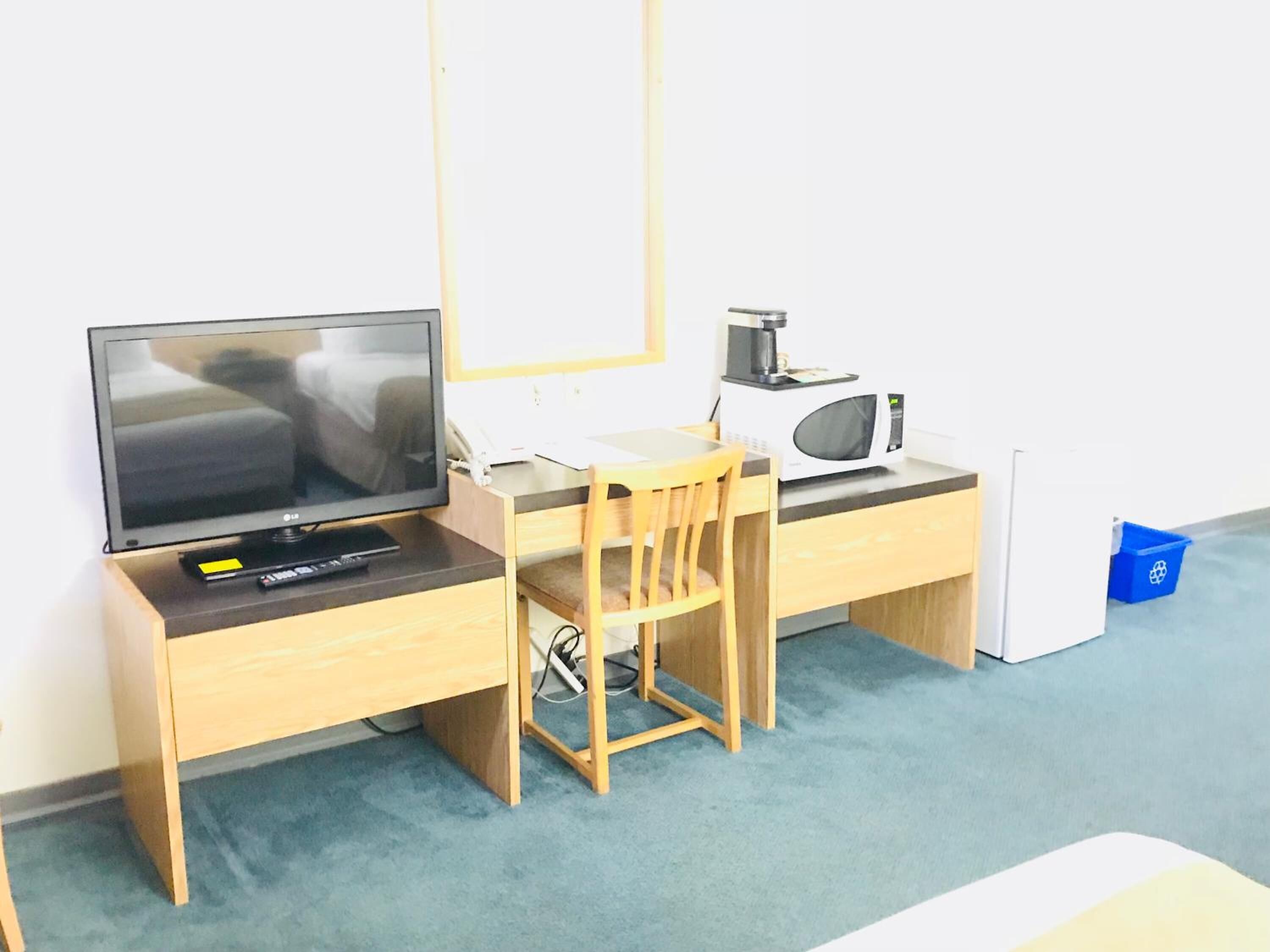 Quarto pequeno e organizado, com uma escrivaninha de madeira, uma cadeira, uma televisão, um micro-ondas e uma pequena lixeira.