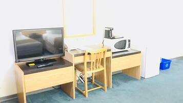 Quarto pequeno e organizado, com uma escrivaninha de madeira, uma cadeira, uma televisão, um micro-ondas e uma pequena lixeira.