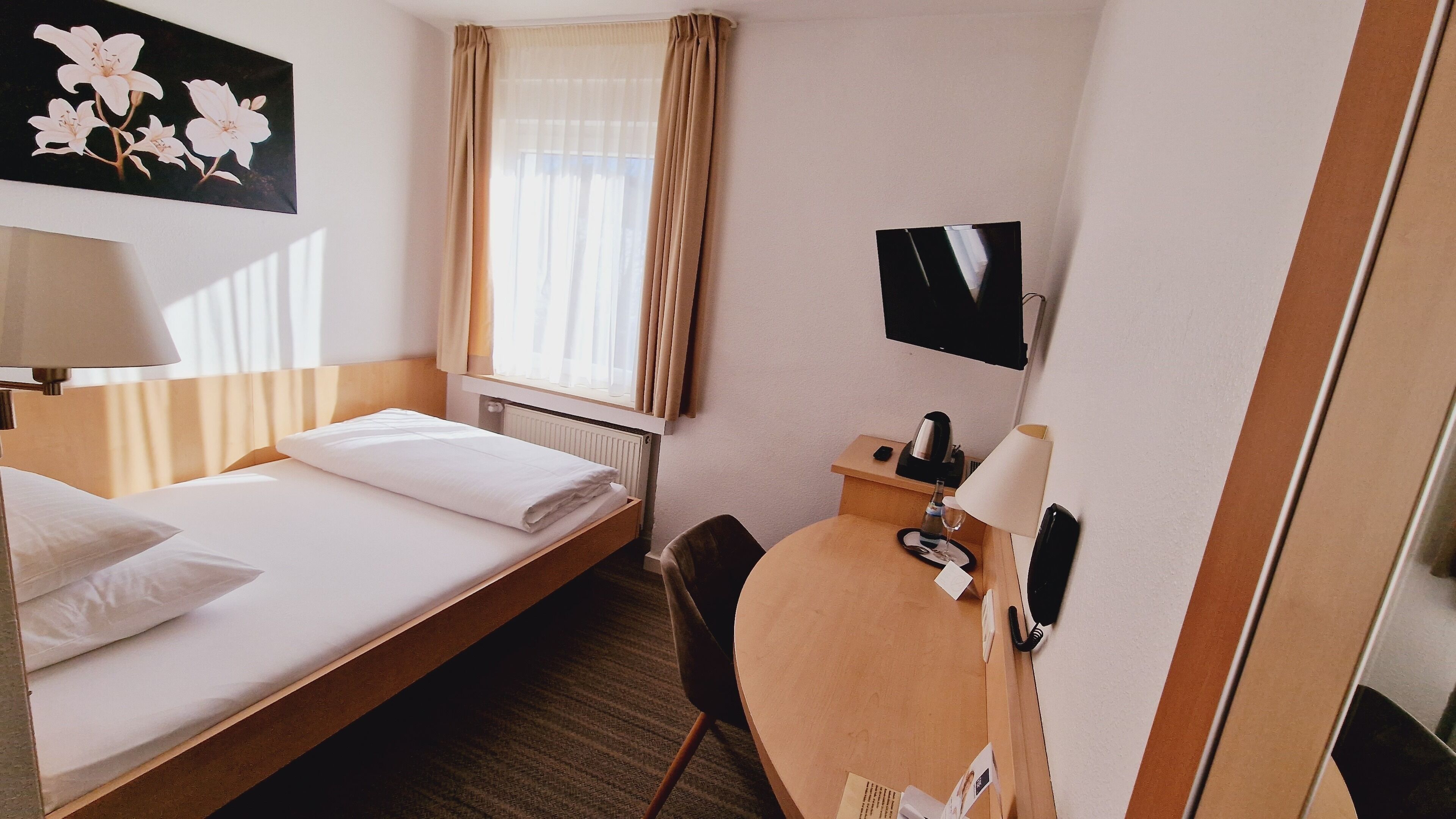 Ein Hotelzimmer mit Bett, Schreibtisch, Stuhl, Lampe, Fernseher und einem Blumenbild.