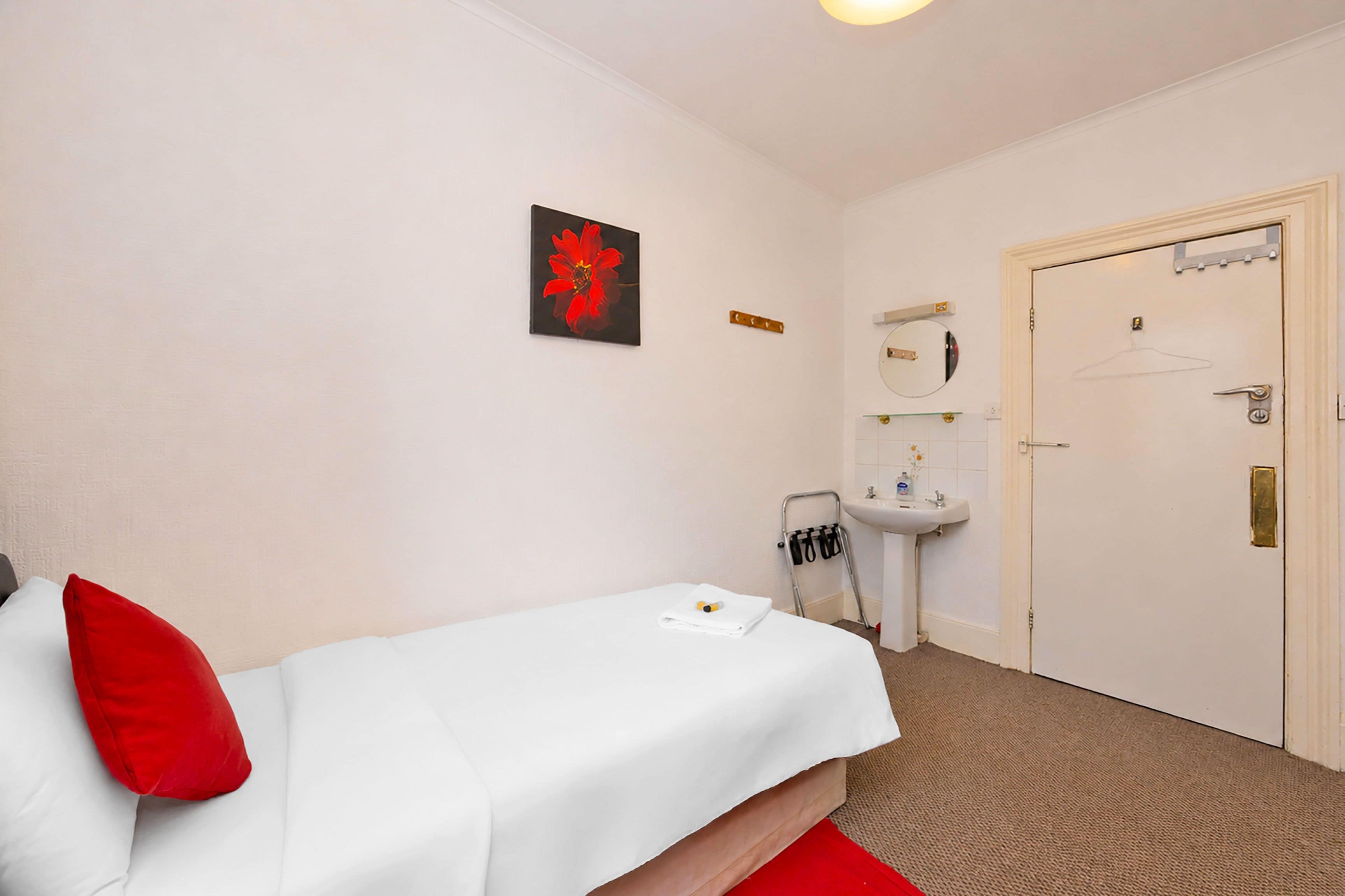 A hotel room with a bed, a red pillow, a white bedspread, a painting of a red flower, a sink, and a door with a hanger.