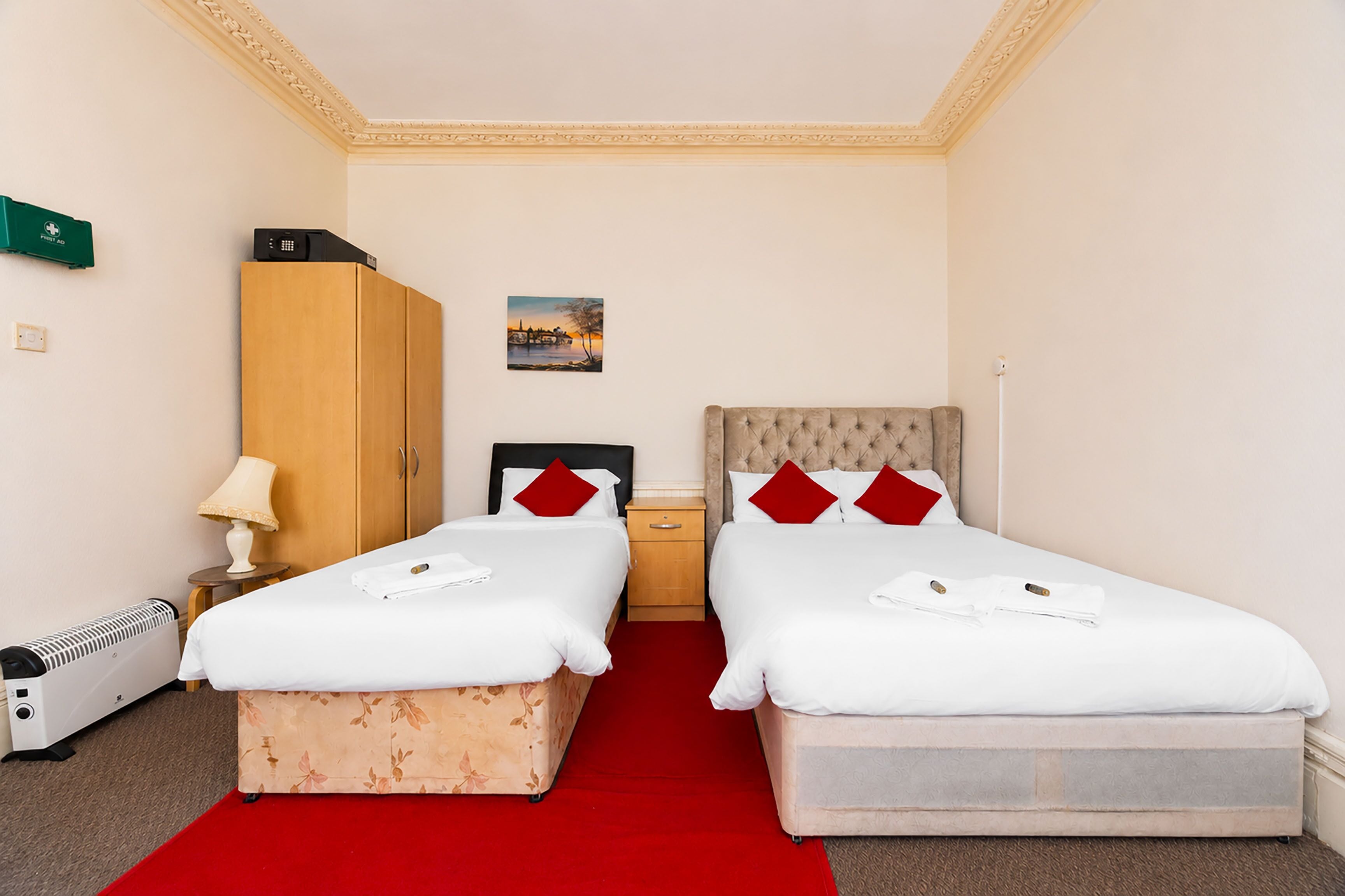 Camera d'albergo con due letti, un armadio in legno, un comodino, una lampada, un termosifone e un piccolo quadro appeso al muro.