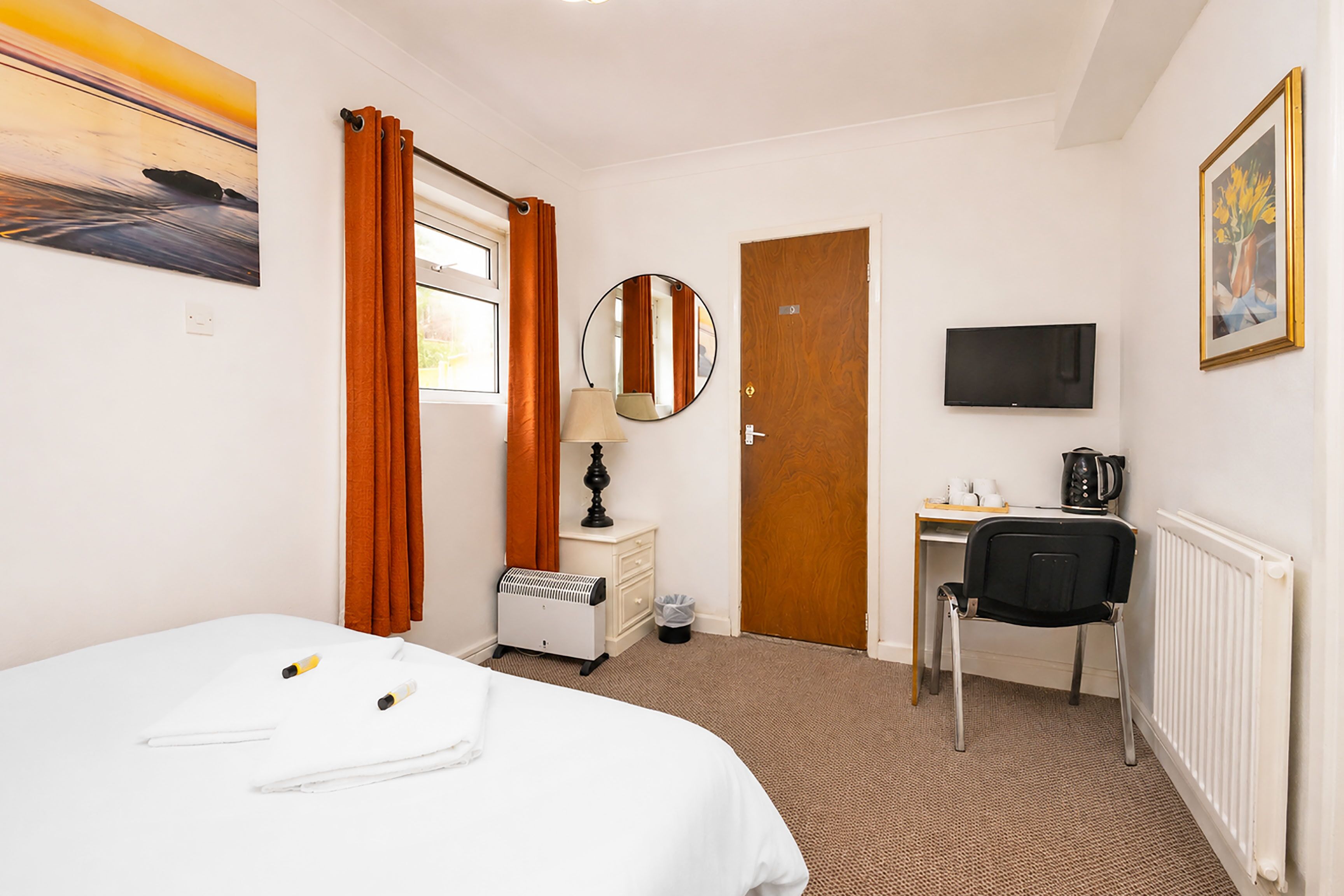 A hotel room with a bed, a desk, a chair, a TV, a radiator, a heater, a mirror, and a window with curtains.