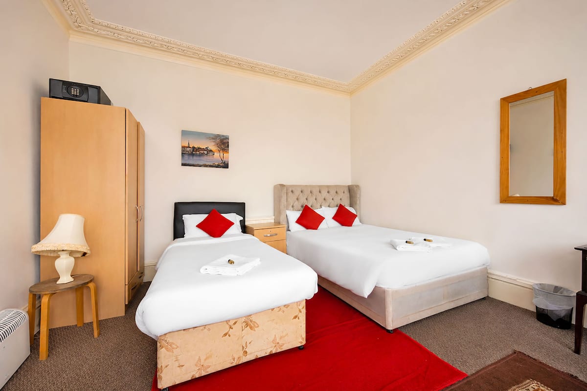 A hotel room with two beds, a wooden wardrobe, a nightstand, a lamp, a painting, and a mirror.