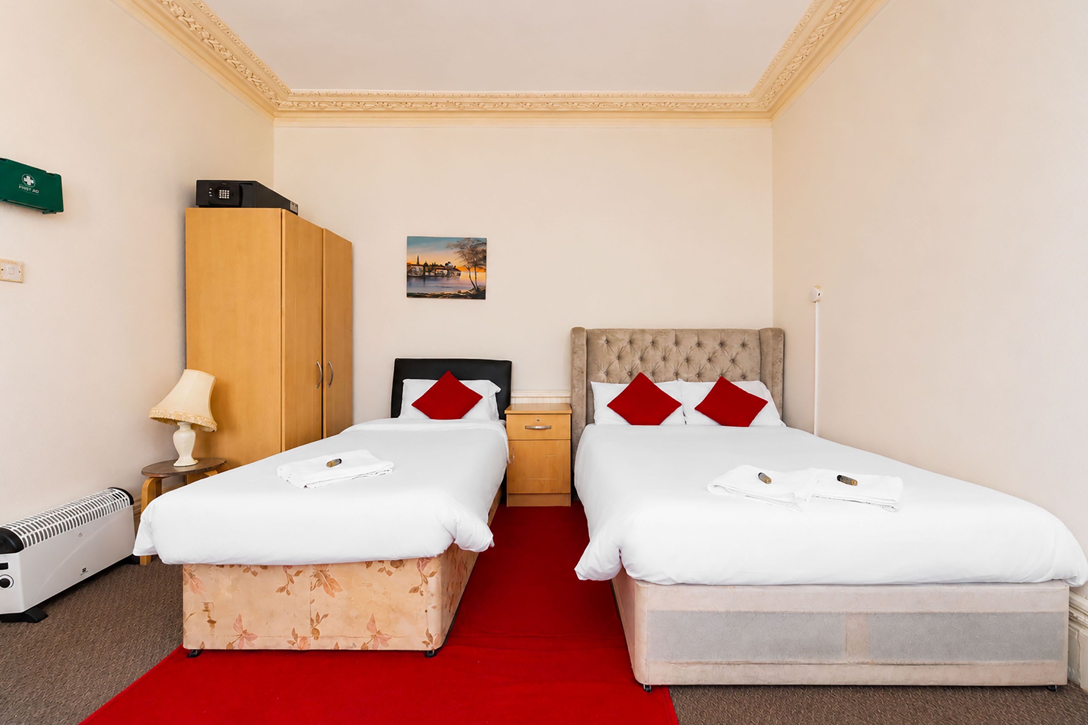 A hotel room with two beds, a red carpet, a wooden wardrobe, a bedside table with a lamp, and a small table with a green object.