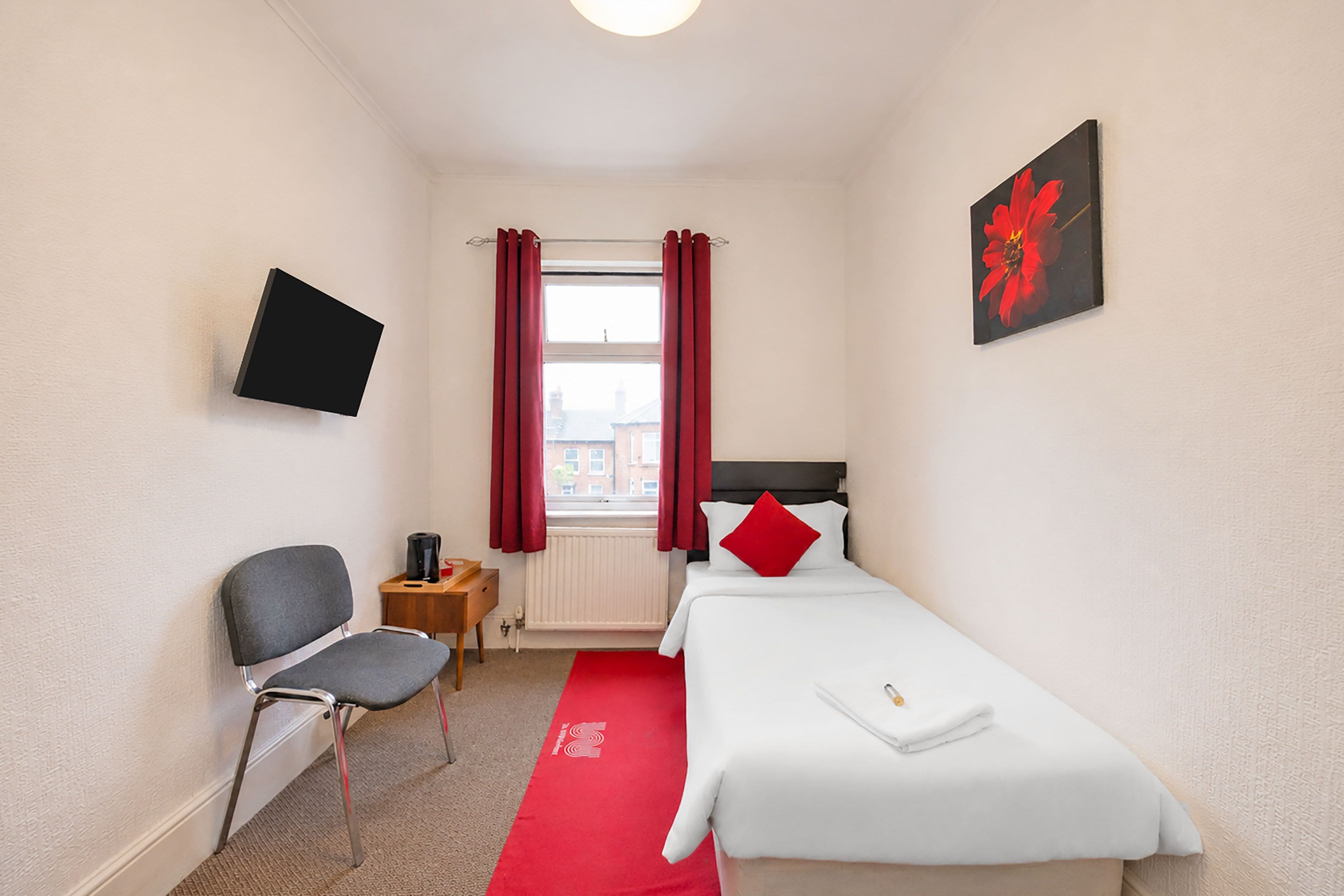 Ein kleines Hotelzimmer mit einem Einzelbett, einem Sessel, einem an der Wand befestigten Fernseher und einem Fenster mit Vorhängen.