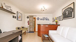 A hotel reception area with a reception desk, a sofa, a coffee table, a desk with a chair, and a wall with framed pictures and a sign. - Saji Hotel Ilford (Ilford)