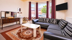 A living room with a sofa, a coffee table, a TV mounted on the wall, a dresser, and a window with curtains. - Saji Hotel Ilford (Ilford)
