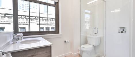 A modern bathroom with a glass shower enclosure, a double sink vanity, and a window providing a view of the cityscape.