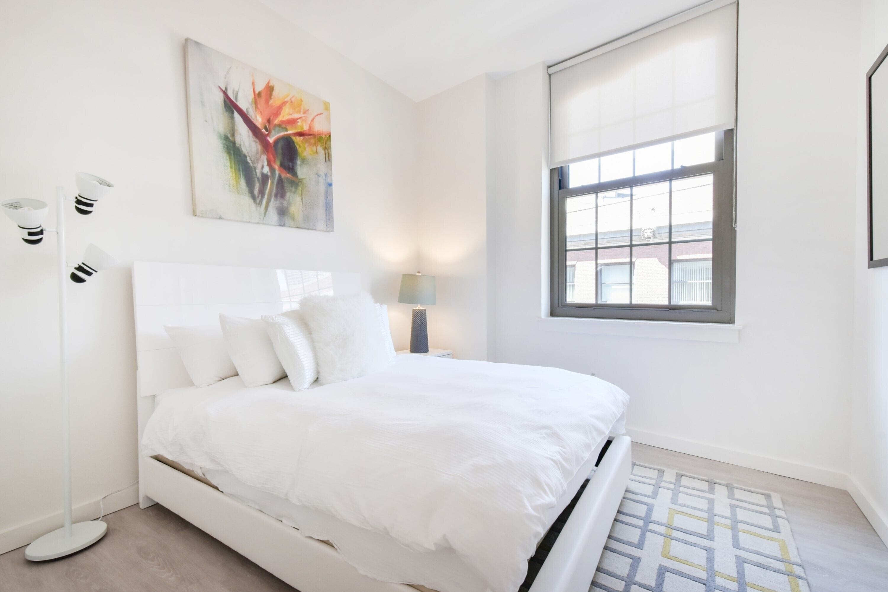 A modern bedroom with a white bed, a floor lamp, a painting, and a large window.
