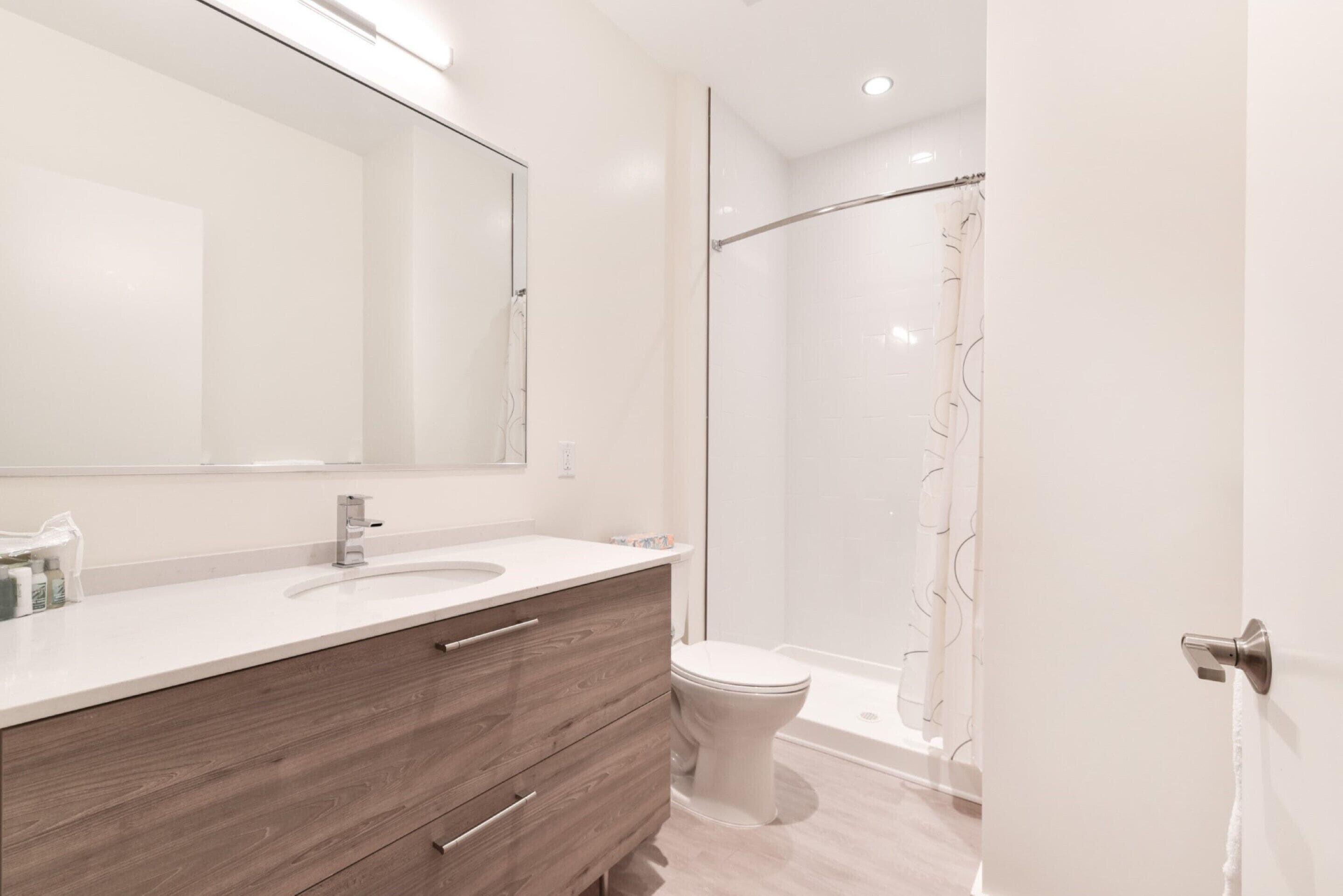 Une salle de bain moderne avec un grand miroir, un meuble-lavabo en bois, un lavabo blanc et un espace douche.