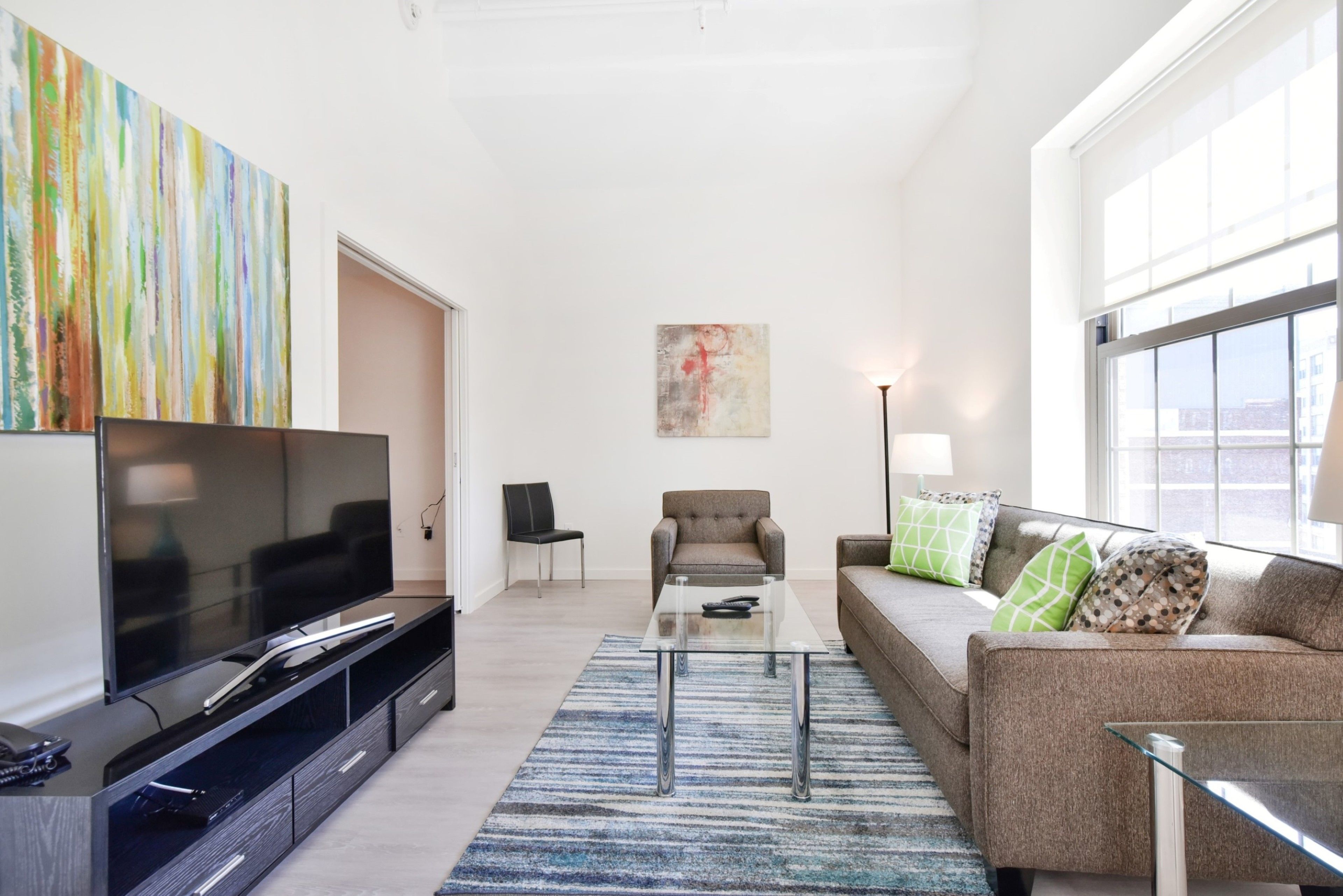 A modern living room with a flat-screen TV, a glass coffee table, a sofa with patterned cushions, and a colorful abstract painting on the wall.