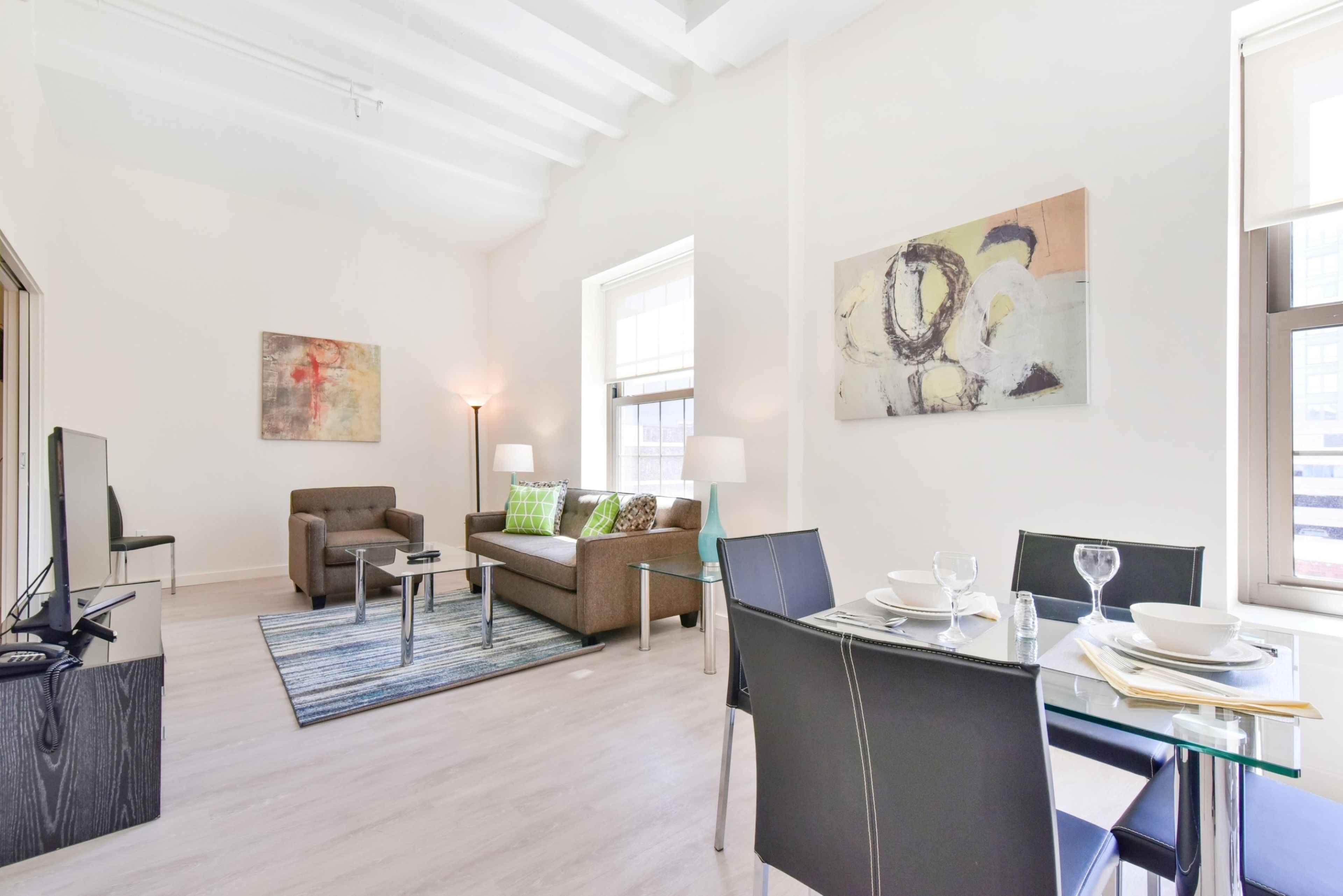 A modern living room with a glass dining table, a sofa, and a coffee table. There is a painting on the wall and a window providing natural light.