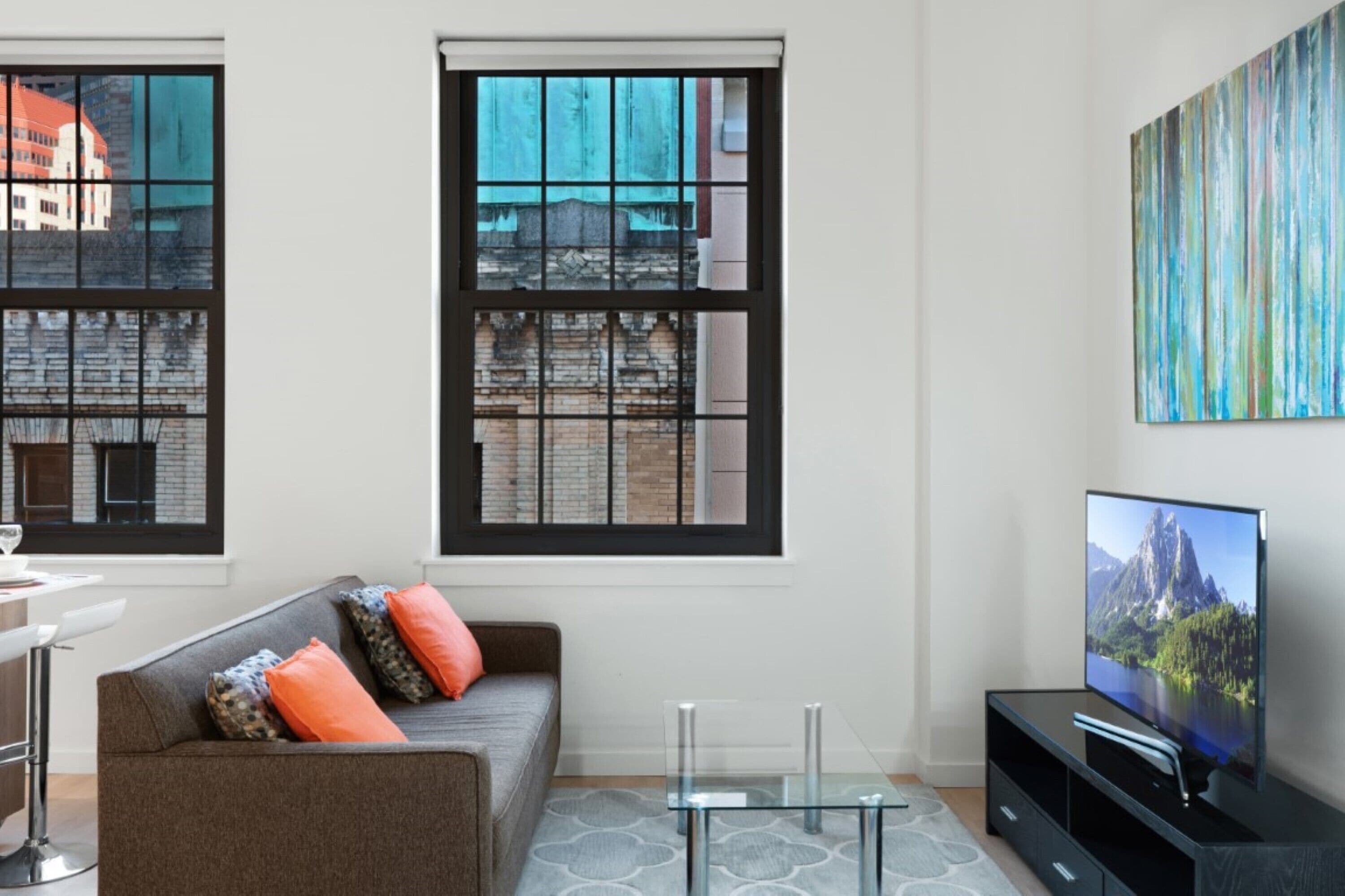 Ein modernes Wohnzimmer mit einer Couch, einem Glastisch, einem Fernseher und großen Fenstern mit Blick auf die Stadt.