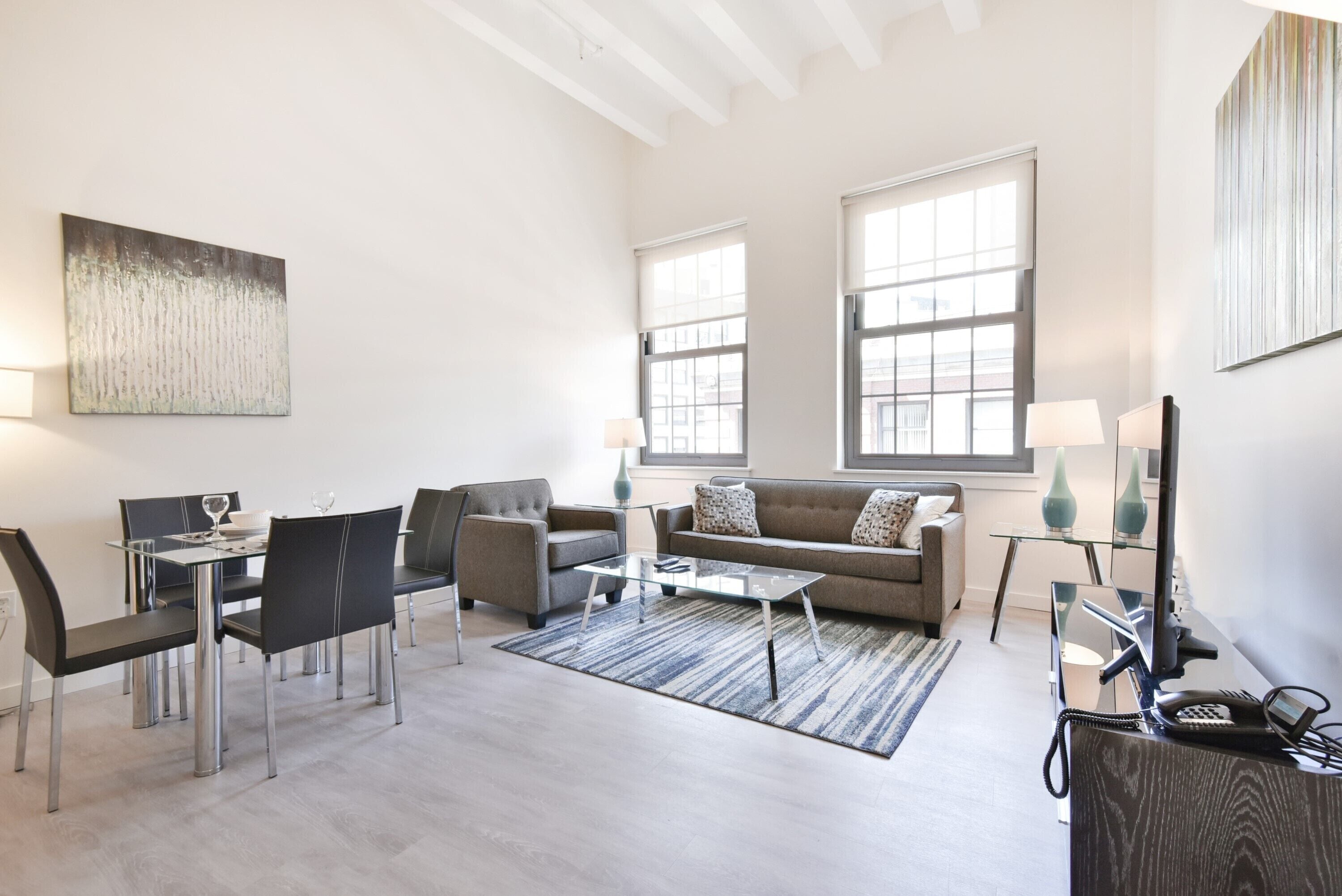 Una sala de estar moderna con sofá, sillones, una mesa de centro y televisor. Amplias ventanas ofrecen luz natural y vistas al exterior.