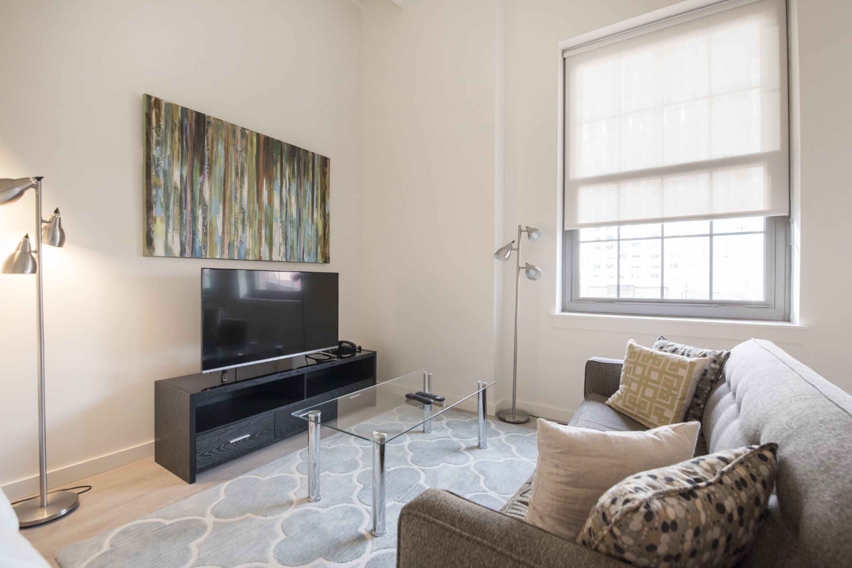 A modern living room with a flat-screen TV, a glass coffee table, a sofa with patterned cushions, a floor lamp, and a large window with blinds.