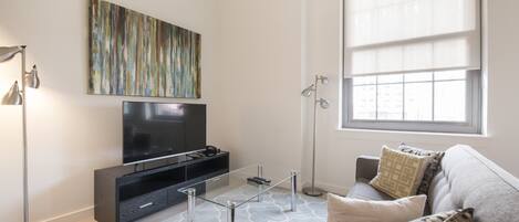 A modern living room with a flat-screen TV, a glass coffee table, a sofa with patterned cushions, a floor lamp, and a large window with blinds.