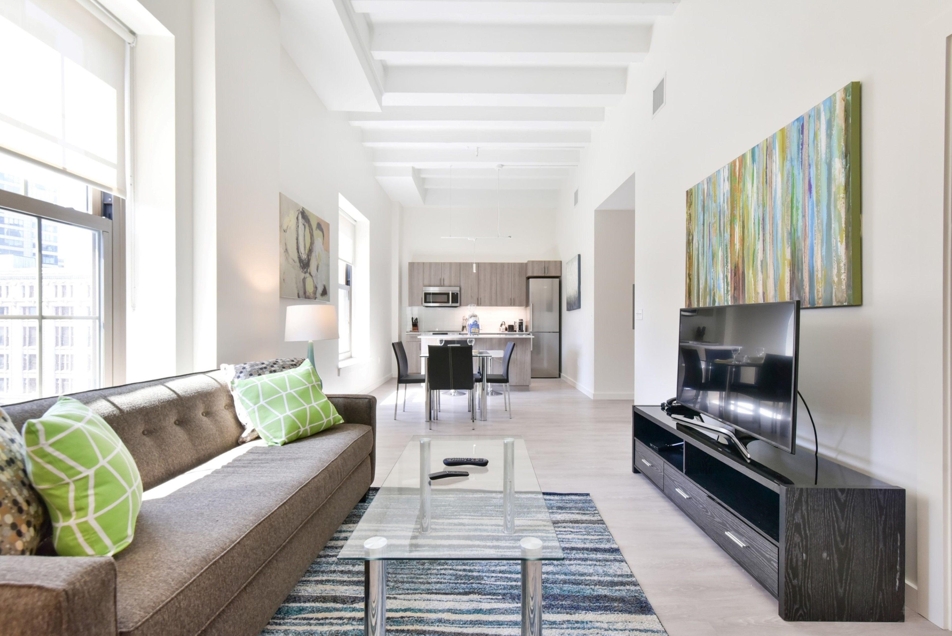 A modern living room with a sofa, a coffee table, and a TV. There is a kitchen in the background with a dining area.