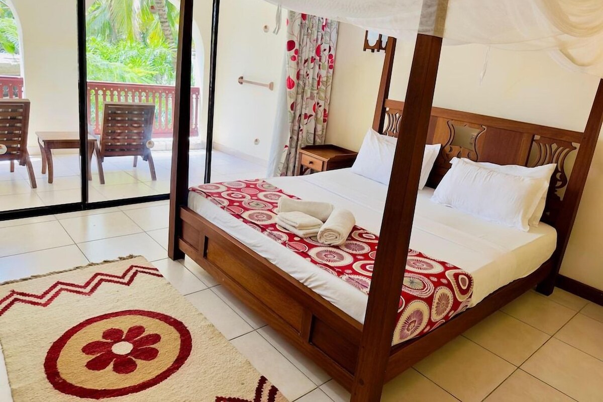 Uma cama com dossel com edredom vermelho e branco estampado, cabeceira de madeira e almofadas brancas. Um pequeno criado-mudo de madeira fica ao lado da cama, com uma luminária. O quarto tem uma janela grande com vista para o verde lá fora.