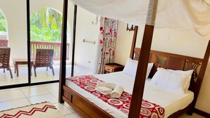 A four-poster bed with a red and white patterned bedspread, a wooden headboard, and white pillows. There is a wooden nightstand with a lamp, a wooden chair, and a small table with a chair outside the room. - Lotfa Resort Diani (Diani Beach)