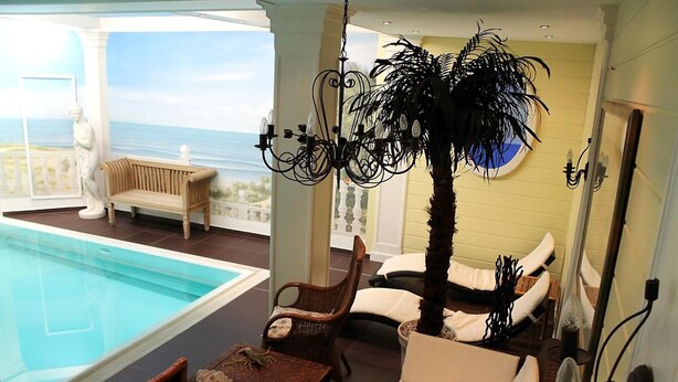 A poolside area with a view of the ocean, a chaise lounge, a dining table, and a decorative palm tree. - Parkhotel Fischer (Wernigerode)