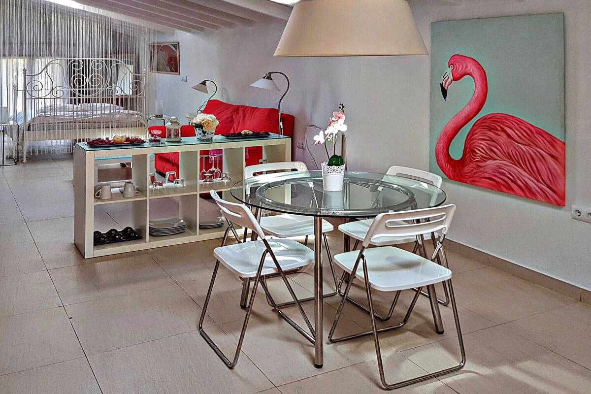 A dining area with a glass table surrounded by white chairs, a red sofa, and a painting of a flamingo on the wall.