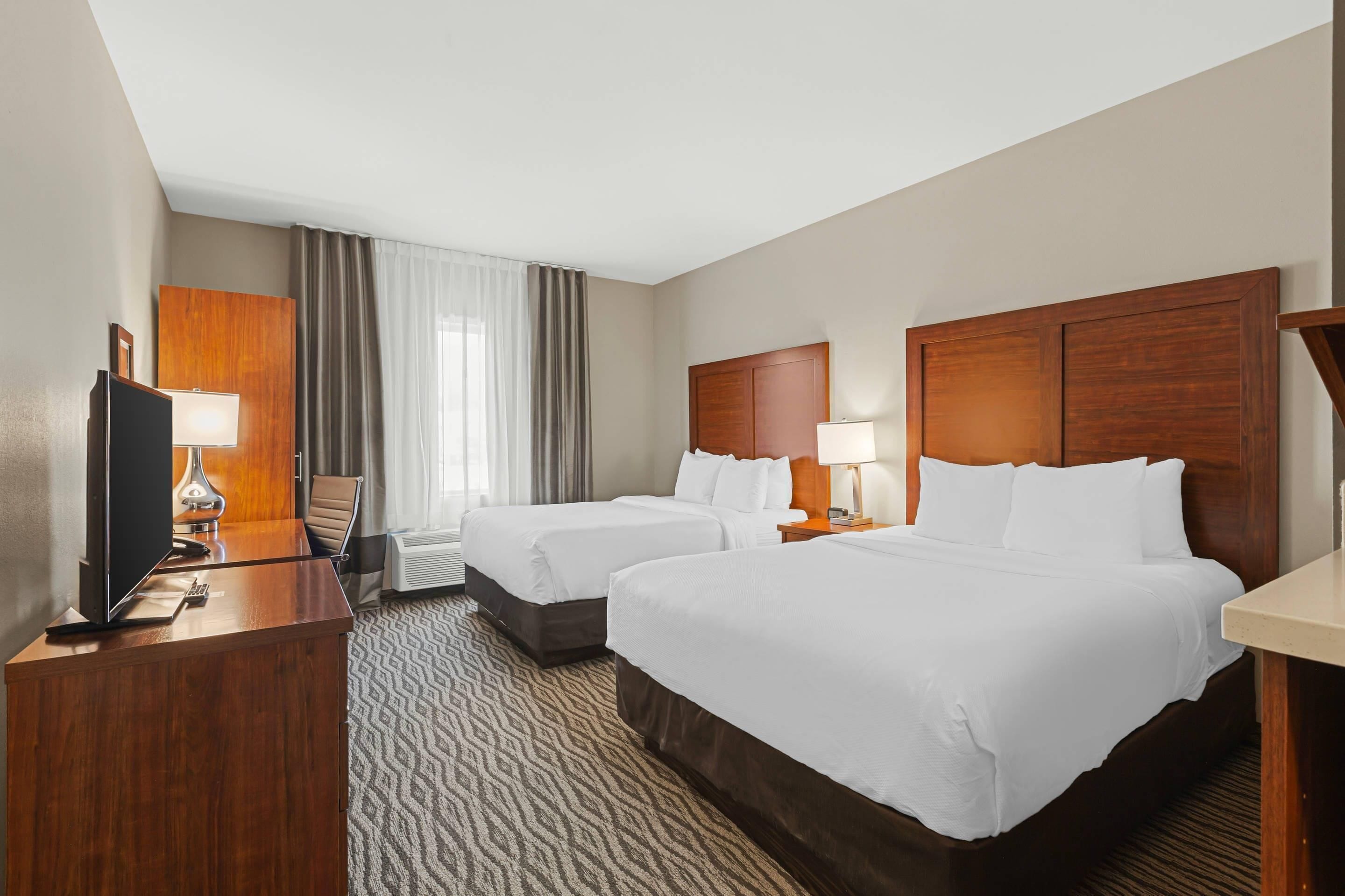 A hotel room with two beds, a desk, a TV, and a large window with curtains.