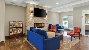 A hotel lobby with a stone wall, a flat-screen TV, a blue sofa, red and blue chairs, a bookshelf, and a patterned rug. - Comfort Inn & Suites (Clarington)
