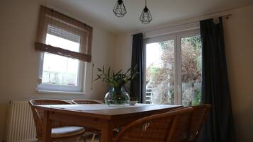 Un comedor con mesa de madera, sillas de mimbre, un florero con plantas y una ventana con persianas.