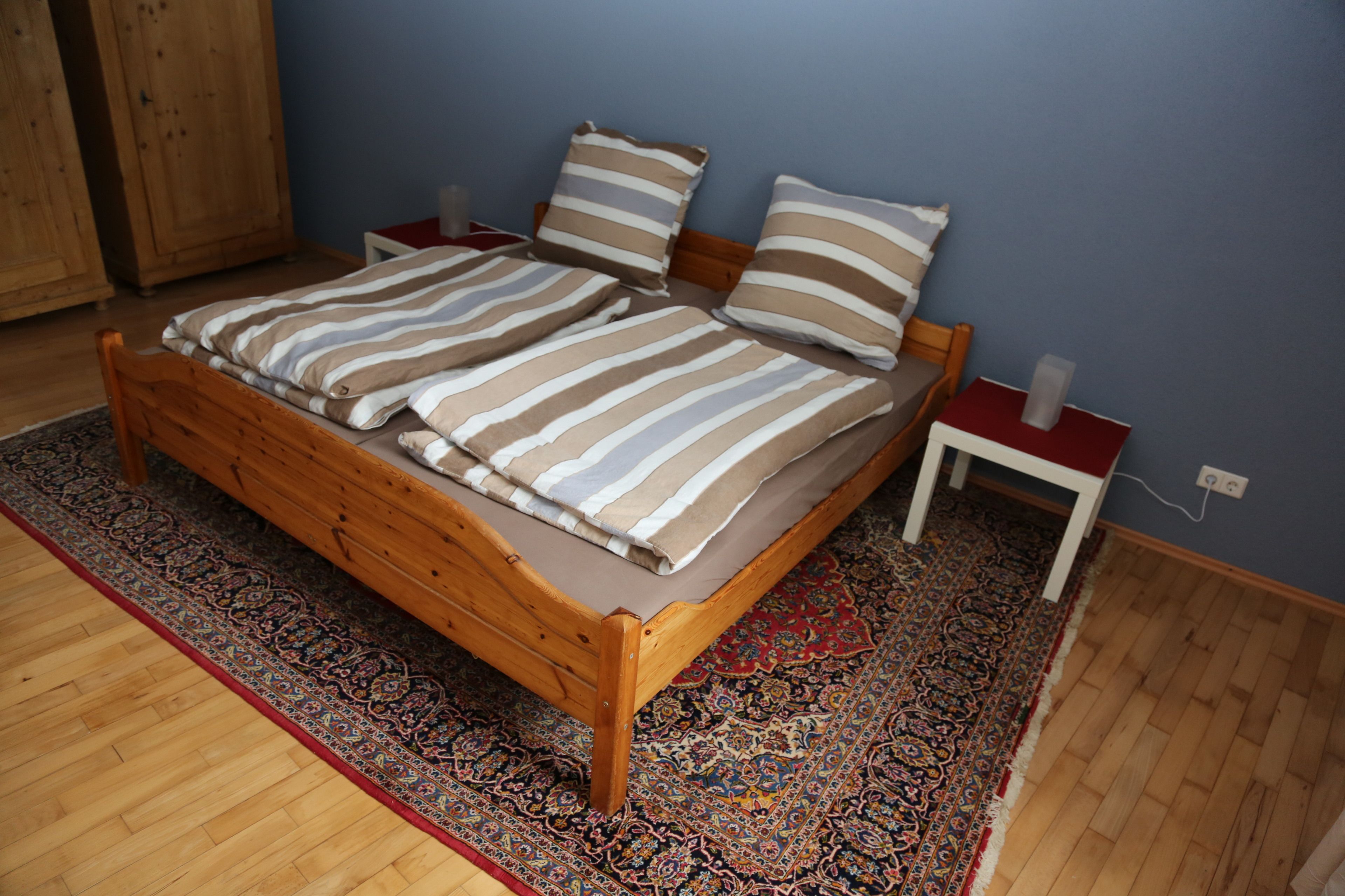 Ein hölzernes Bett mit gestreifter Bettwäsche, ein Teppich mit blau-rotem Muster und ein kleiner Nachttisch mit rotem Kissen.