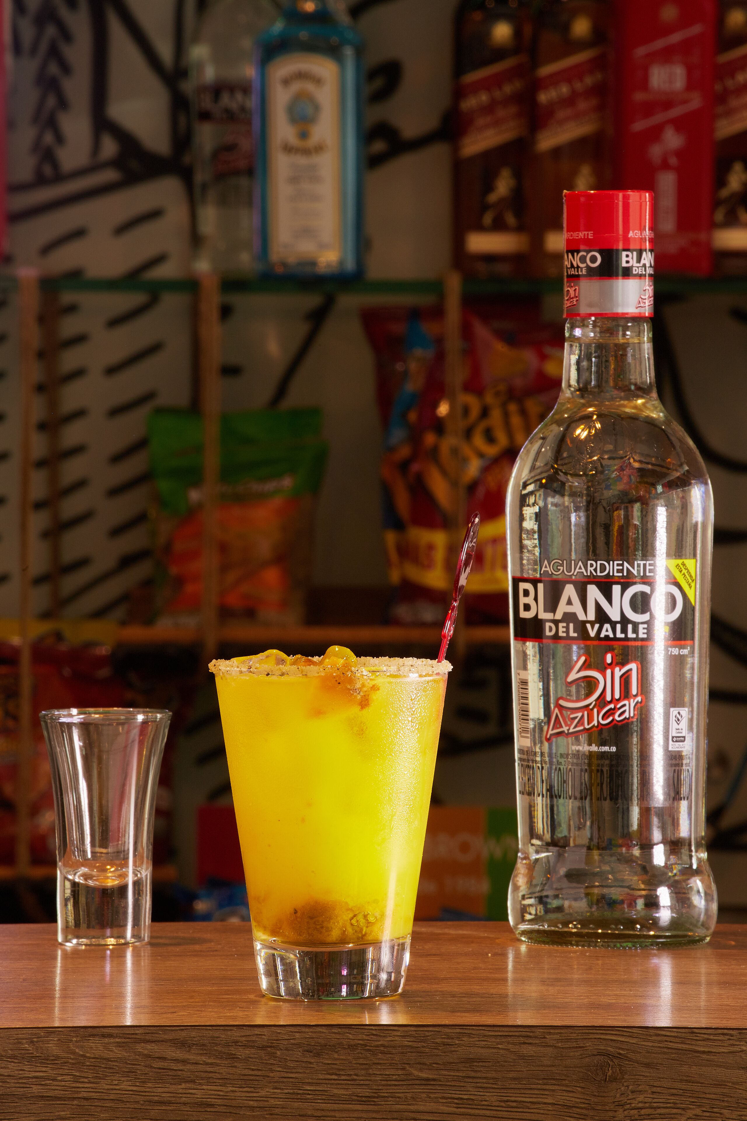 A bottle of Blanco del Valle 3 in Sugar tequila, a glass with a yellow drink, and a small glass of water on a wooden surface.