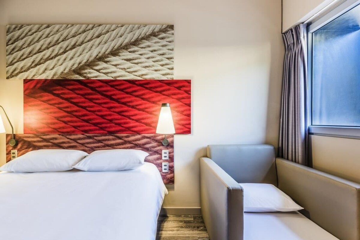 A hotel room with a bed, a sofa, a wall-mounted lamp, and a window with curtains.