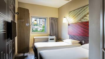 A hotel room with two beds, a TV, a window with a view of buildings, and a lamp.