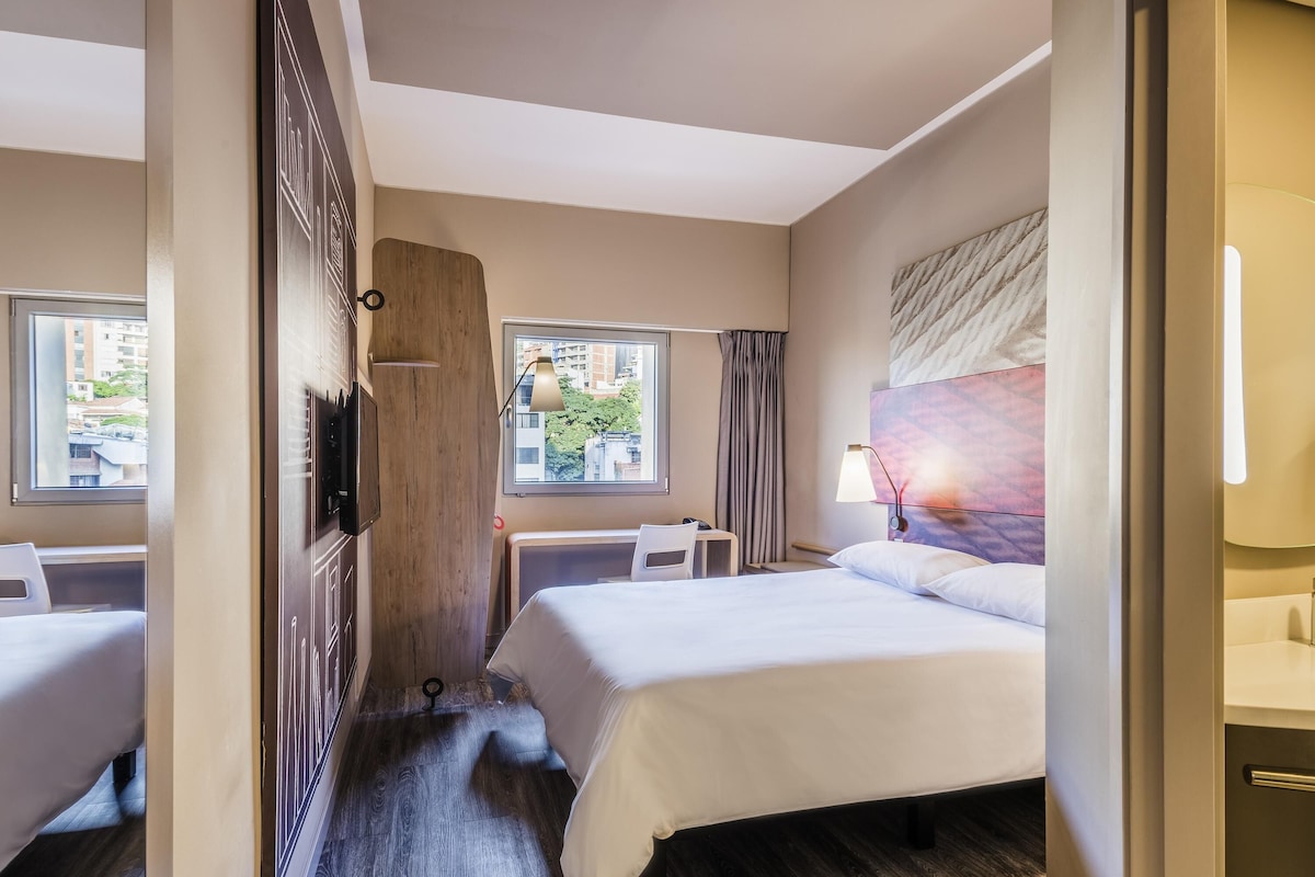 A hotel room with two beds, a desk, a chair, a TV, a window with a view of buildings, and a bathroom with a mirror and sink.