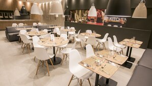 Un restaurant moderne avec des tables rondes et des chaises blanches, agrémenté de luminaires suspendus et d’une grande fresque murale.