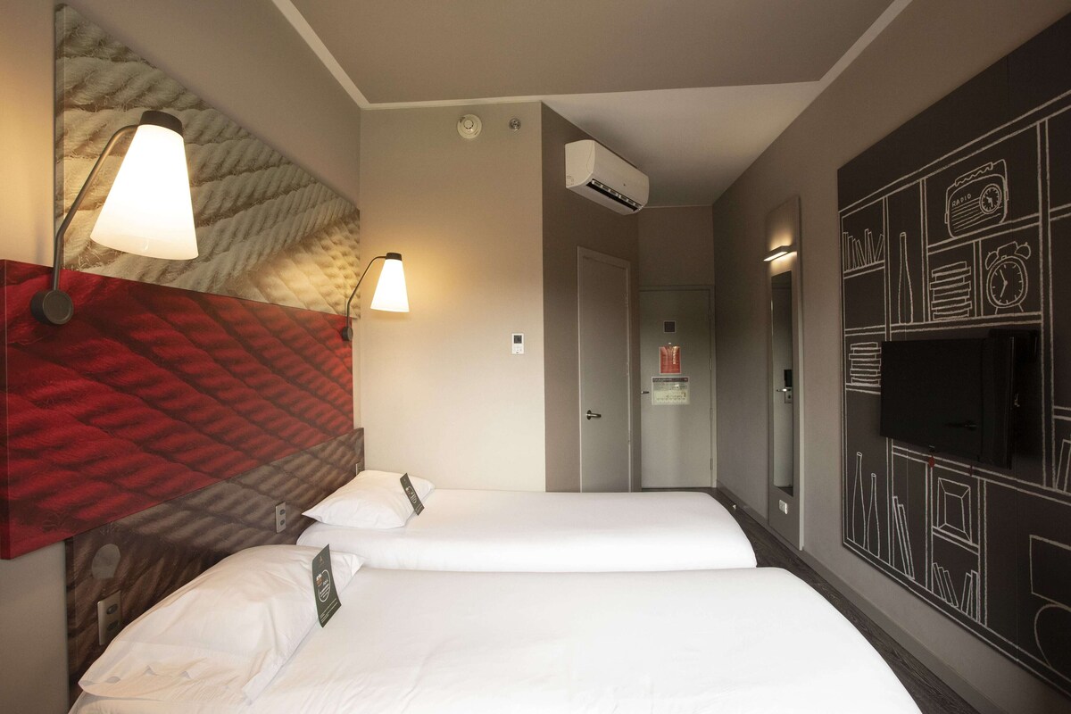 A hotel room with two beds, a wall-mounted TV, an air conditioning unit, and a patterned headboard.
