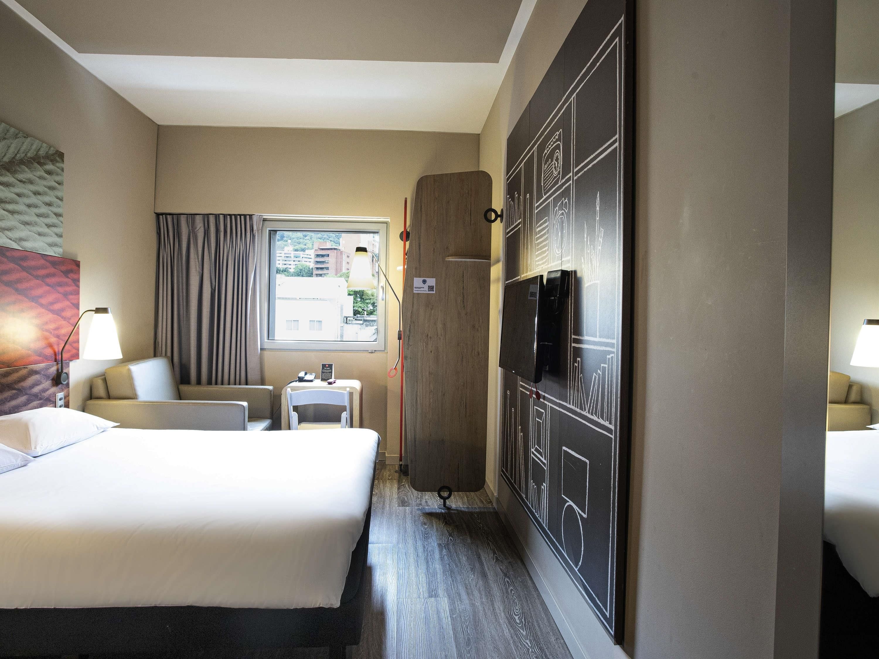 A hotel room with a bed, a desk, a chair, a TV, and a window with curtains.