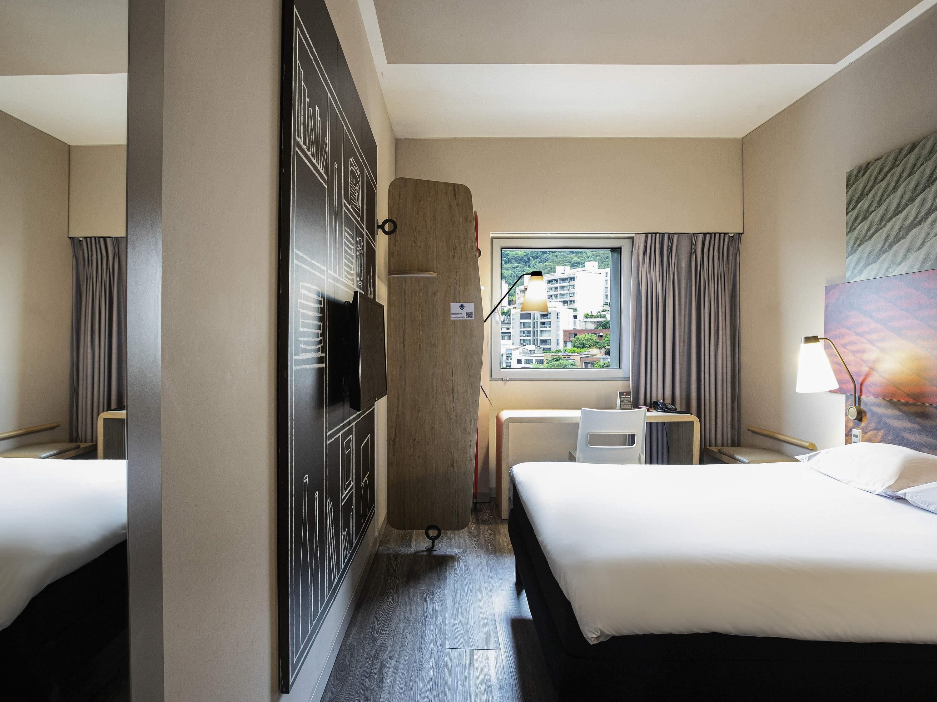 A hotel room with two beds, a desk, a chair, a TV, a window with a view of buildings, and a lamp.
