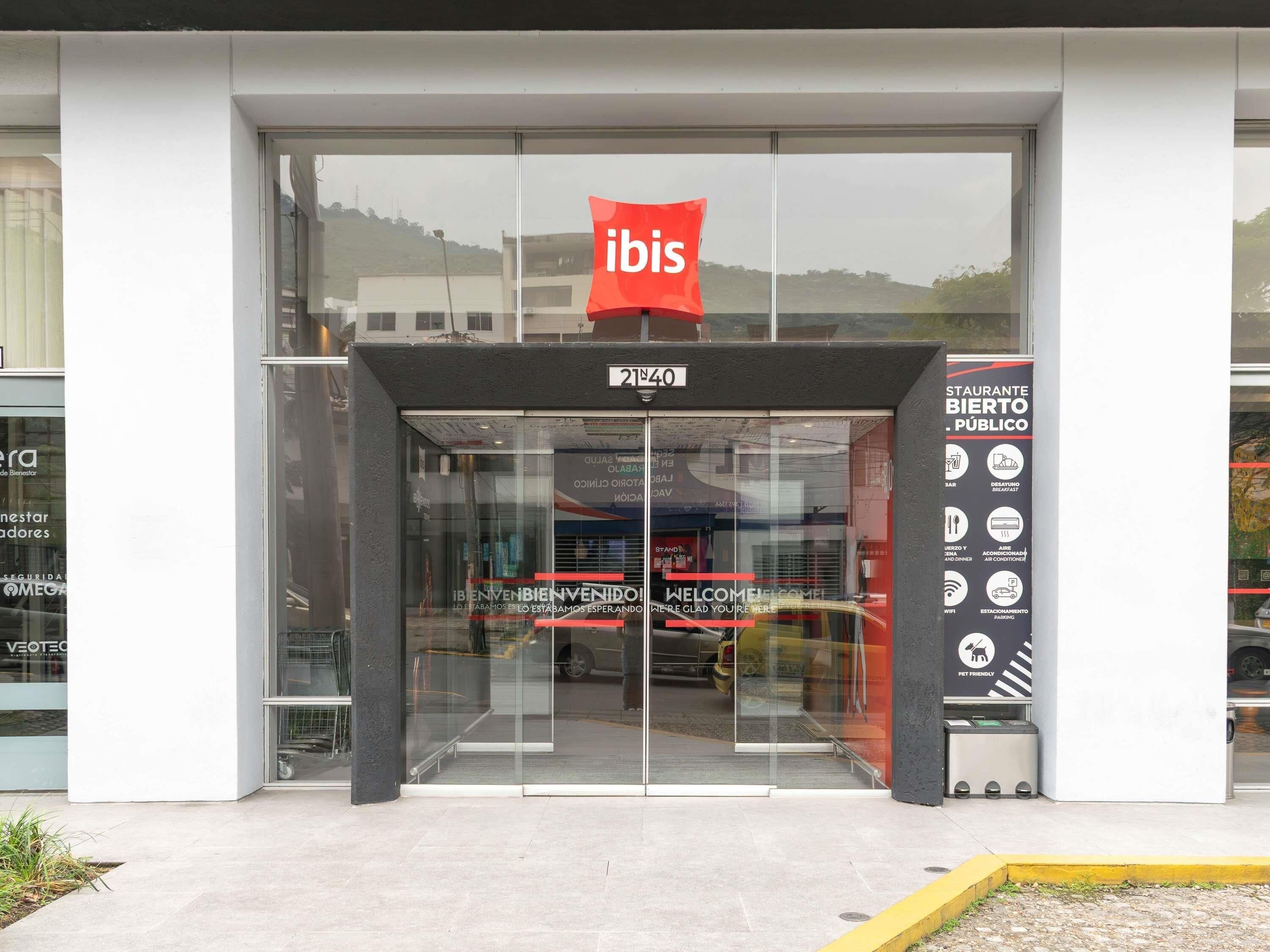 A modern hotel entrance with a glass facade, a red 'ibis' sign, and a welcome banner.