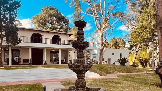 Una fuente con elaboradas esculturas en el centro de un patio, rodeado de un césped cuidado y un edificio con fachada blanca y ventanas arqueadas.
