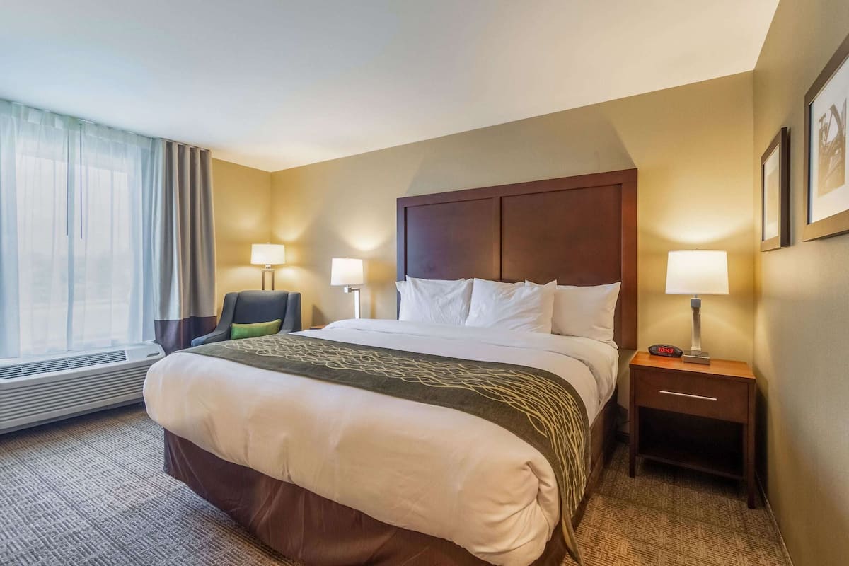 A hotel room with a large bed, two bedside tables with lamps, a chair, a window with curtains, and a wall-mounted picture.