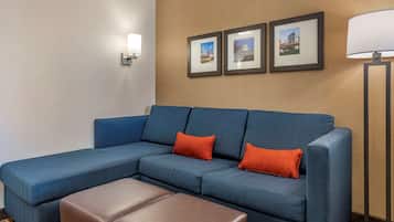 A blue sectional sofa with orange cushions, a brown ottoman, and framed pictures on the wall.