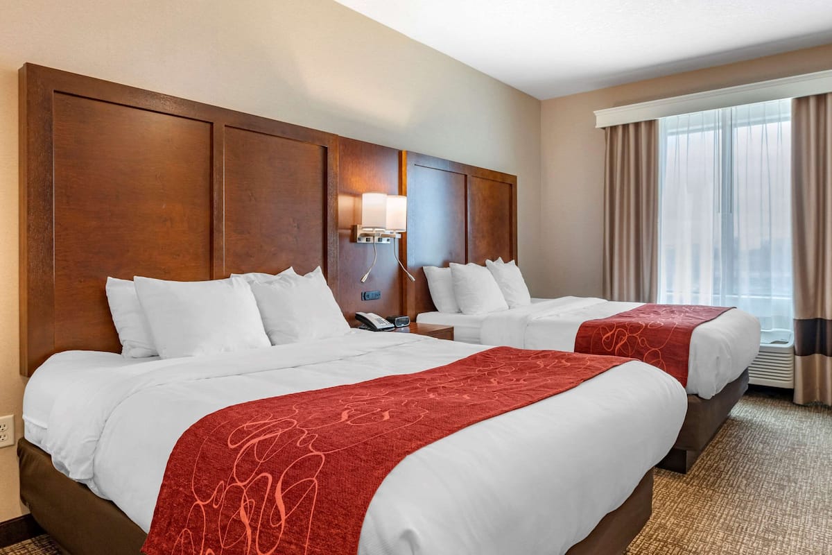 A hotel room with two beds, a wooden headboard, a bedside lamp, and a window with curtains.