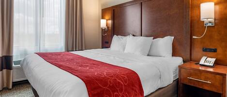 Ein Hotelzimmer mit einem großen Bett, einem hölzernen Kopfteil, einem Nachttisch mit Telefon und einem fensterbelaubten Fenster.