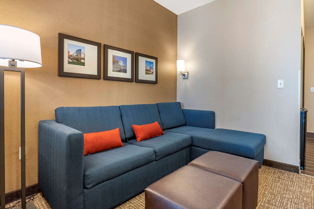 A hotel room with a sectional sofa, two red throw pillows, a brown ottoman, a floor lamp, and three framed pictures on the wall.