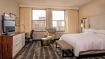 A hotel room with a bed, a desk with a TV, a chair, a lamp, and a view of buildings through large windows.