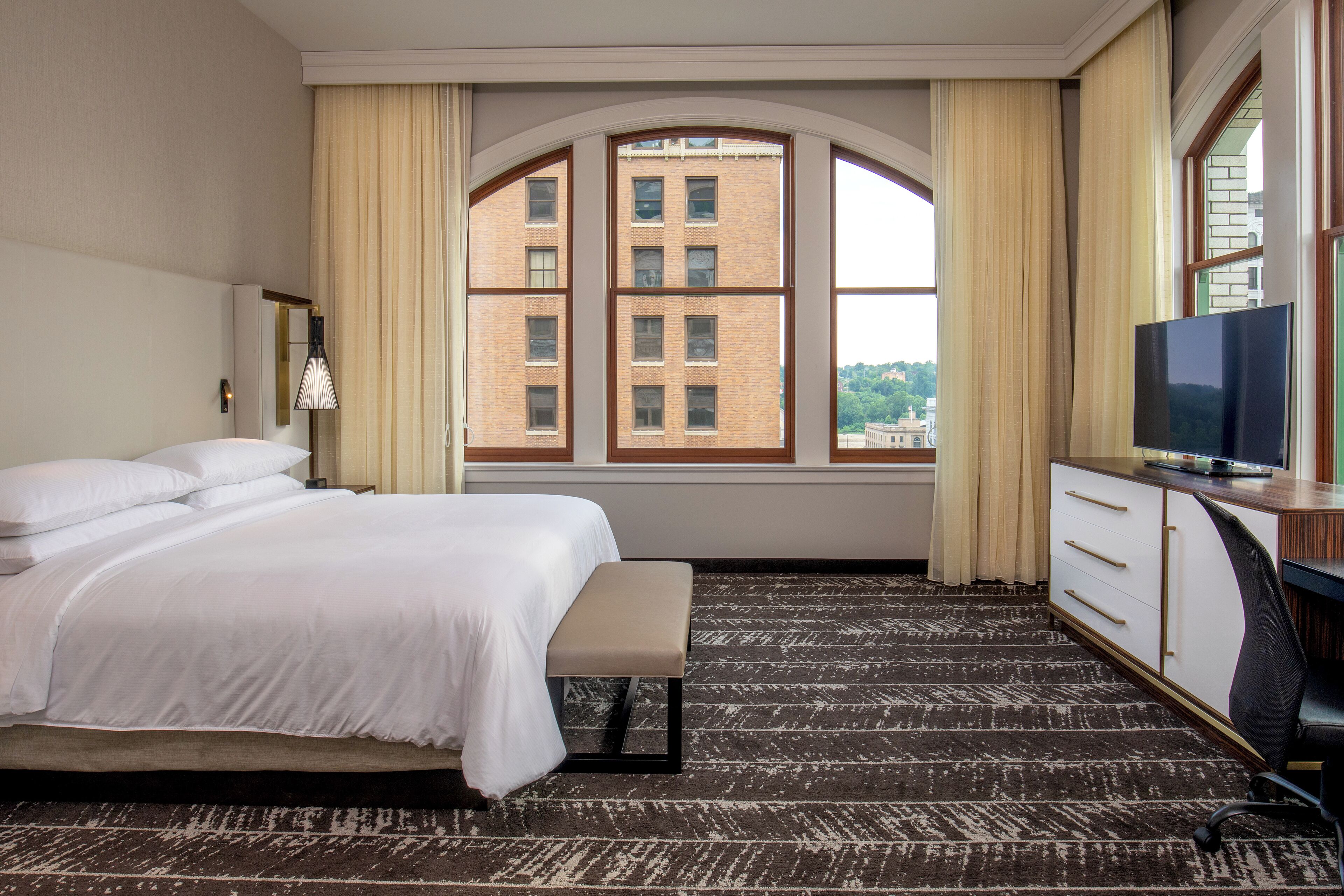 A hotel room with a bed, a desk, a TV, and a large window with curtains.