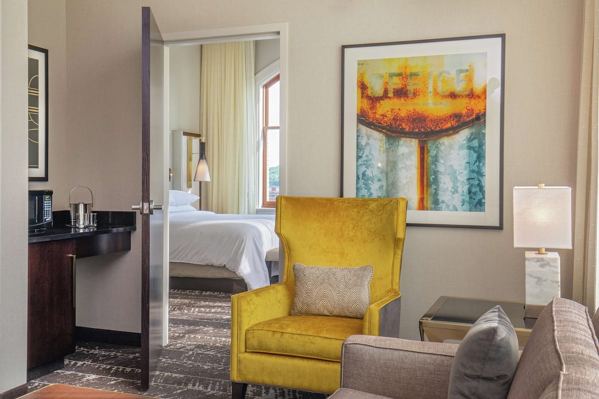 Ein Hotelzimmer mit gelbem Sessel, grauem Sofa, Bett, Schreibtisch und einem Bild an der Wand.