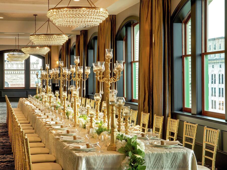 Uma sala de banquetes com longas mesas preparadas para um evento formal, com lustres e grandes janelas com vista para edifícios.