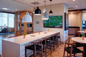 A modern bar area with a long counter, multiple bar stools, pendant lighting, and a television displaying a sports game. - Fairfield Inn & Suites by Marriott Columbus, IN (Columbus)