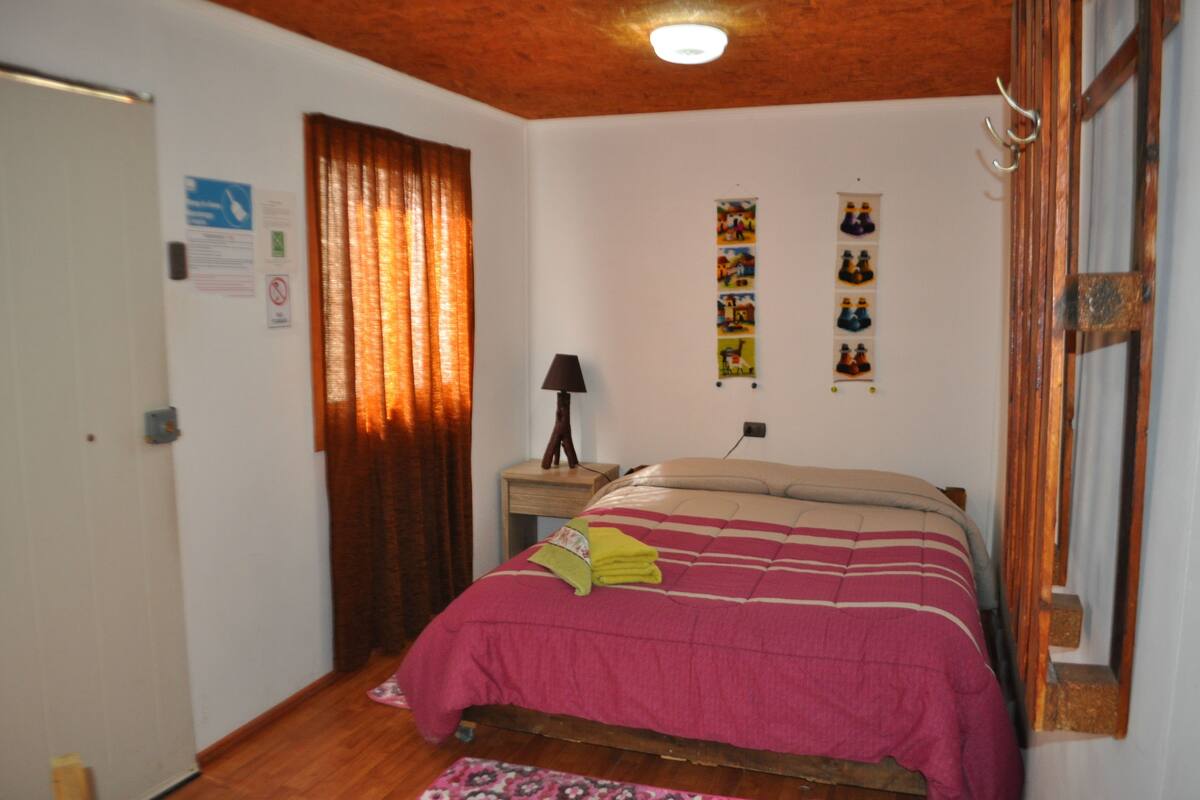 Une chambre à coucher avec un lit, une table de chevet, une lampe, une fenêtre avec des rideaux et un mur décoré de tableaux.